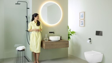 Woman in yellow dress standing in front of mint green bathroom with furniture and bathroom ceramics from Geberit and black taps